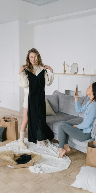 Two women trying on dresses in a stylish living room, surrounded by shopping bags and cozy decor.